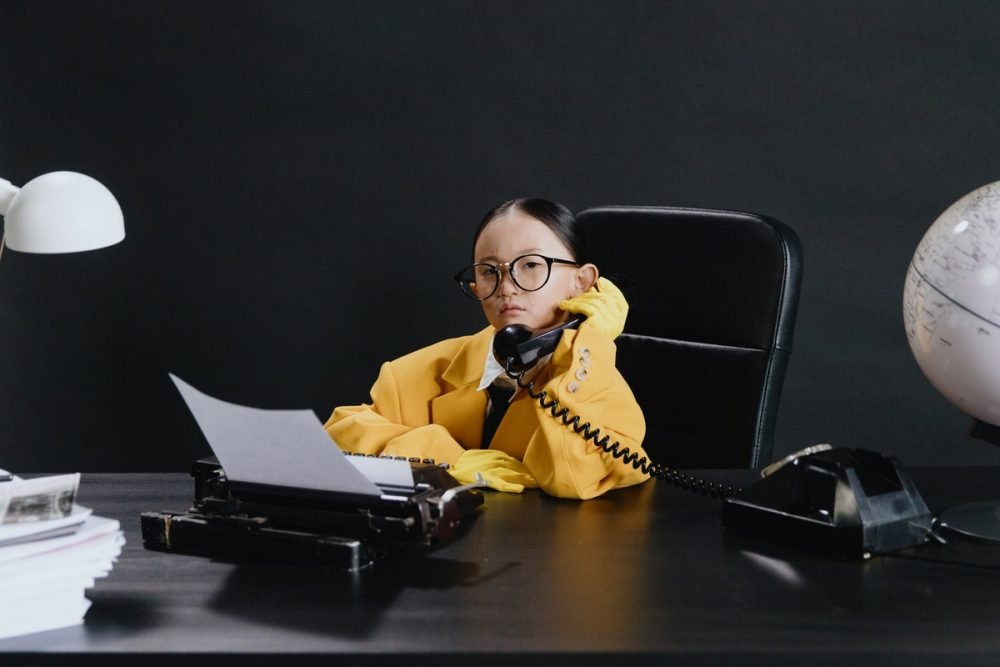 图片:穿着一套成人大小的衣服的女孩,坐在办公桌和拿着电话接收器。