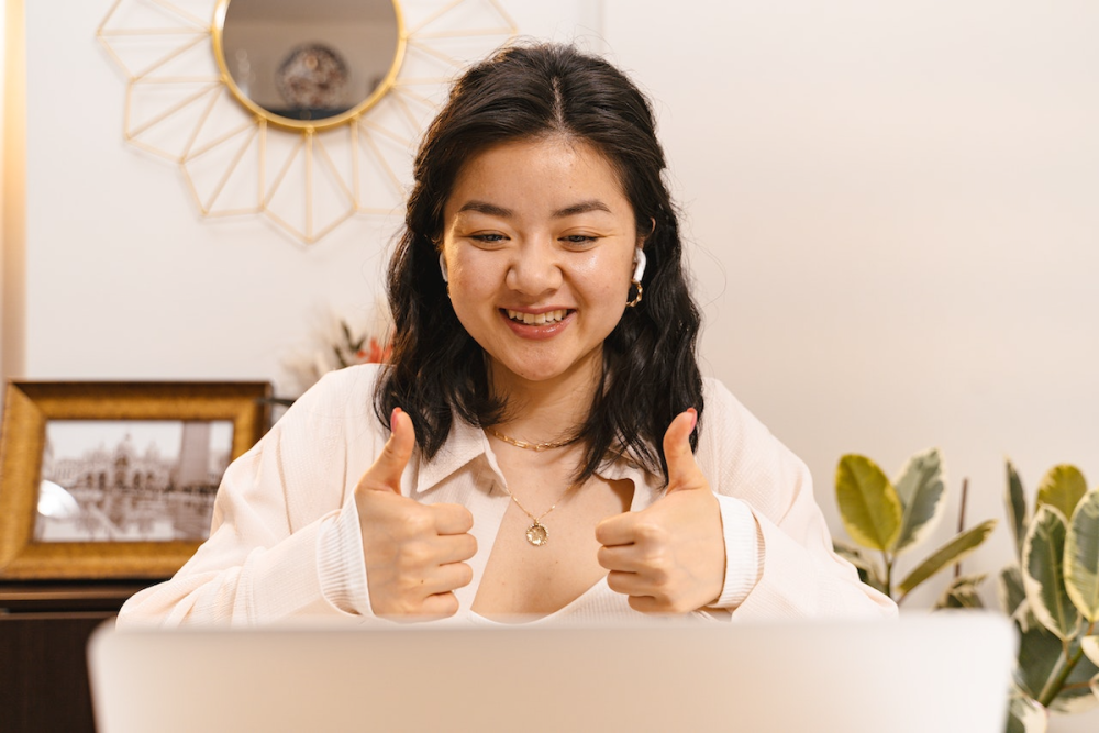 图片:坐在笔记本电脑前,一个微笑的女人戴着耳塞给两个大拇指到电脑屏幕上。
