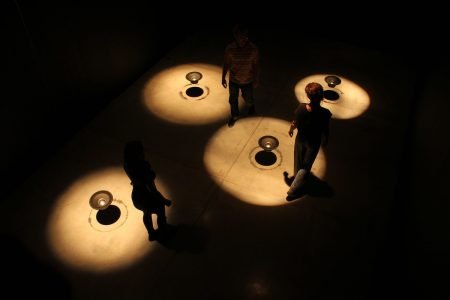 图片：艺术安装暗室，包含四个音频扬声器，悬挂在中空中，由聚光灯点亮，人们走过空间。