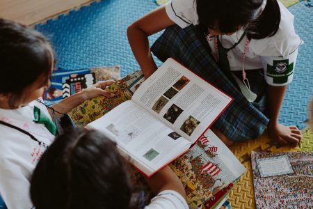 图片：三个女学生聚集在一堆图画书周围。