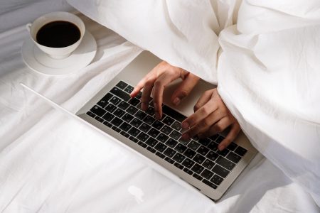 图片：从床床罩上涌出的手在笔记本电脑上打字，床单附近有一杯咖啡。