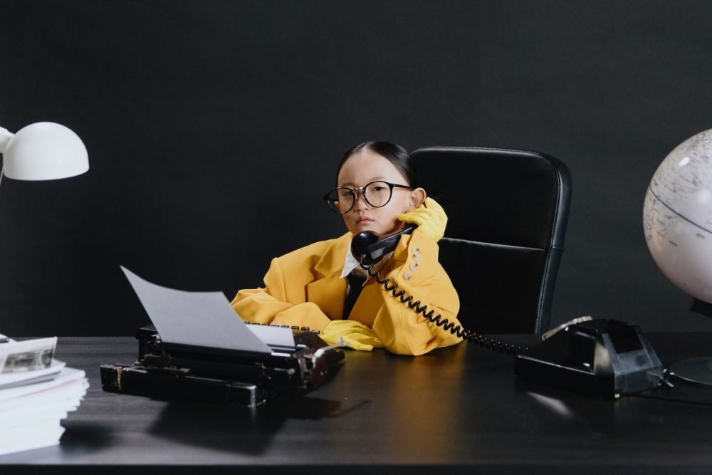 图片：穿着一套成人大小的衣服的女孩，坐在办公桌和拿着电话接收器。