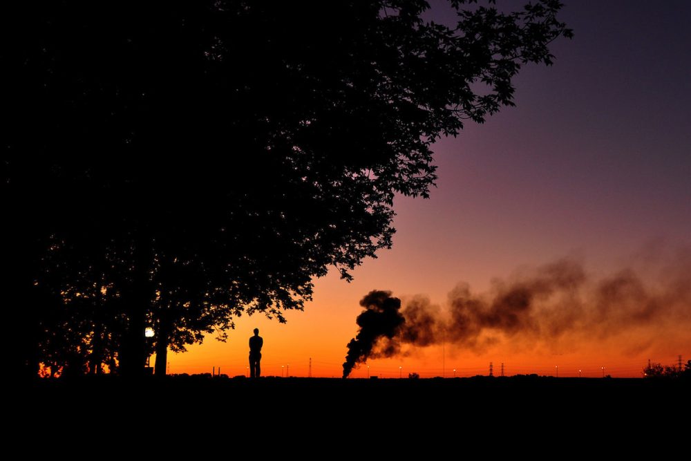 图片：一个孤独的人站着看着遥远的烟雾升起，在五颜六色的日出时剪影。