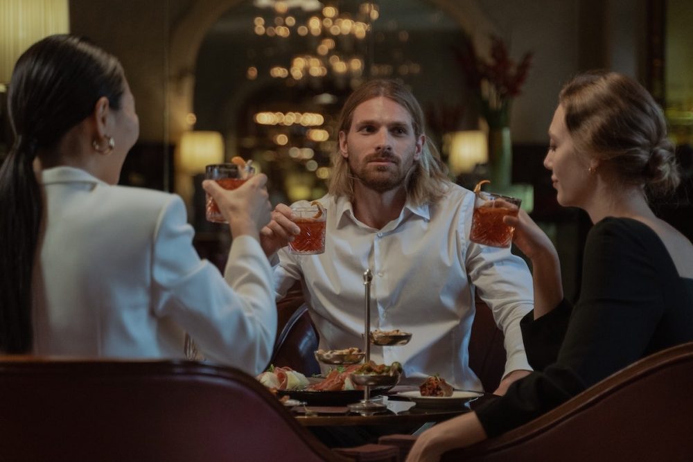 图片:一名男子和两名女子手拿鸡尾酒，围坐在餐厅的桌子旁。