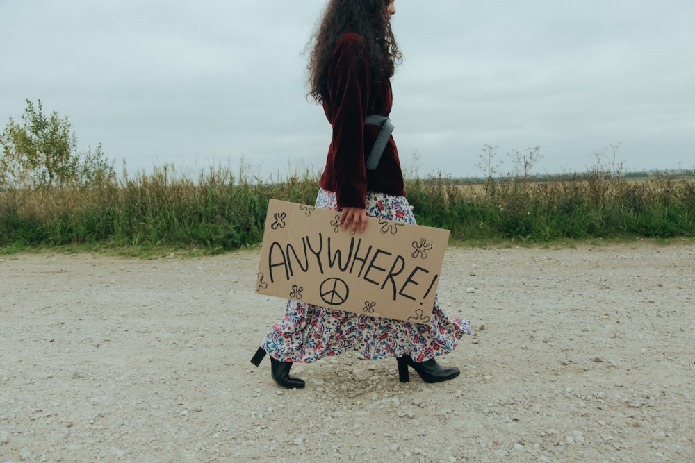 图片:一名身穿波西米亚服饰的搭便车女子，走在土路上，手举手工纸板牌子，上面写着“任何地方!”