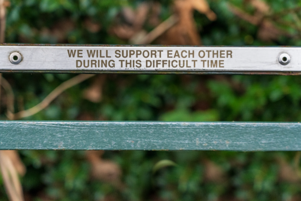 图片:公园长椅后面的一条板条上有一块牌子，上面写着“在这段困难时期，我们将相互支持。”