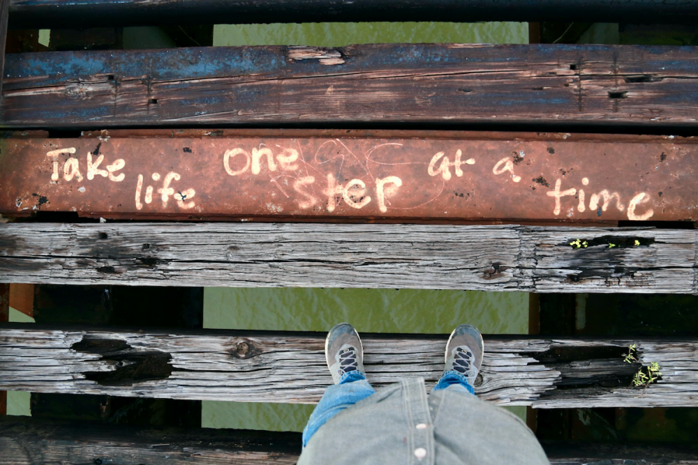 图片:一个人站在废弃人行桥的腐烂木板上。木板之间有很大的缝隙，透过缝隙可以看到水。一块木板上写着“生活一步一个脚印”。