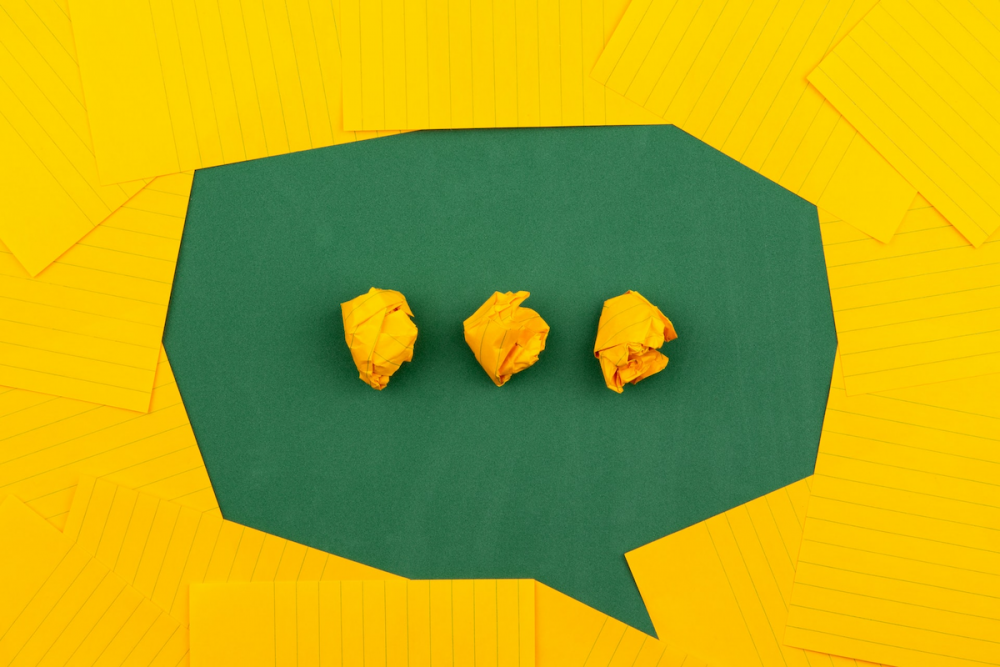 图片:黄色的便签纸被安排在绿色背景上，代表卡通语音气泡的形状，三团皱巴巴的黄色纸在中心像一个省略号一样排列。