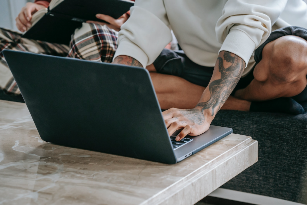 图片:一个身上有大量纹身的男人穿着便服盘腿坐在沙发上，在笔记本电脑上打字，而另一个人坐在他旁边看书。