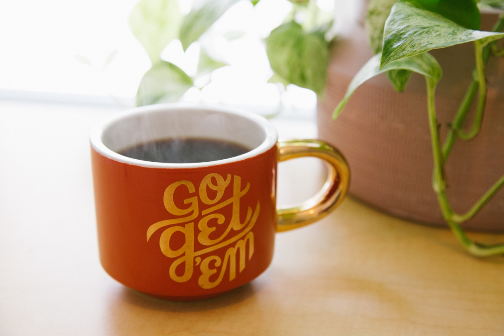 图片:在一盆盆栽和阳光明媚的窗户旁边，有一杯冒着热气的咖啡，上面写着“去拿吧”。