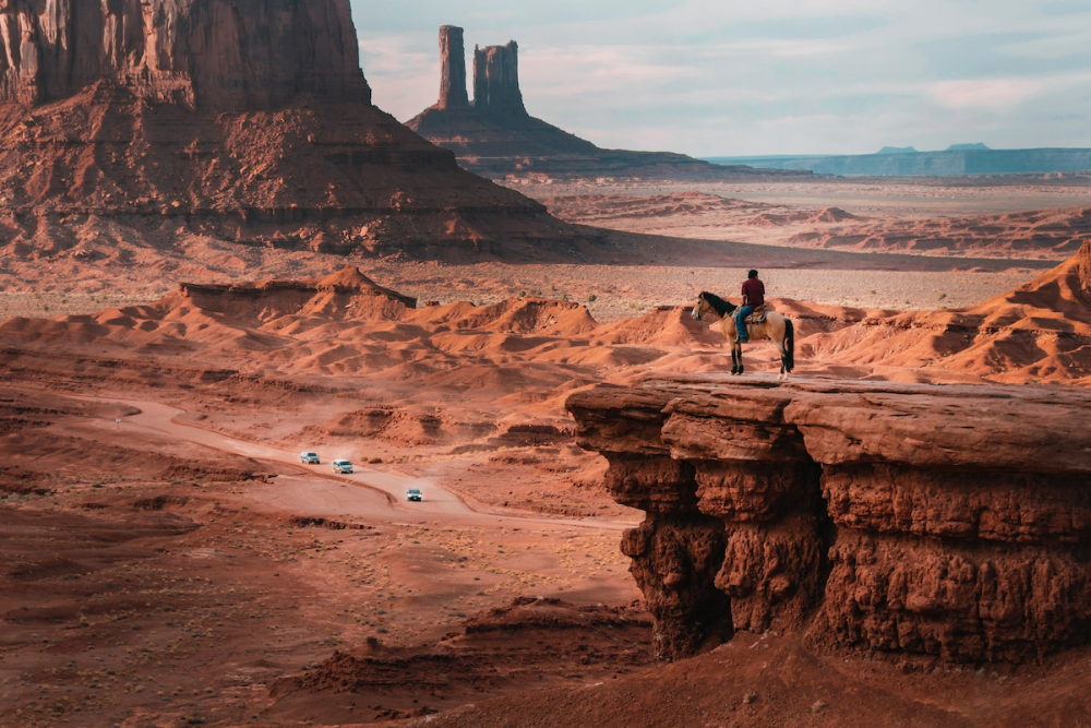 一名穿着便装的美国原住民男子跨骑在悬崖上，俯瞰亚利桑那州奥加托纪念碑谷的景观。在下面的道路上，两辆货车和一辆汽车沿着蜿蜒的道路行驶。