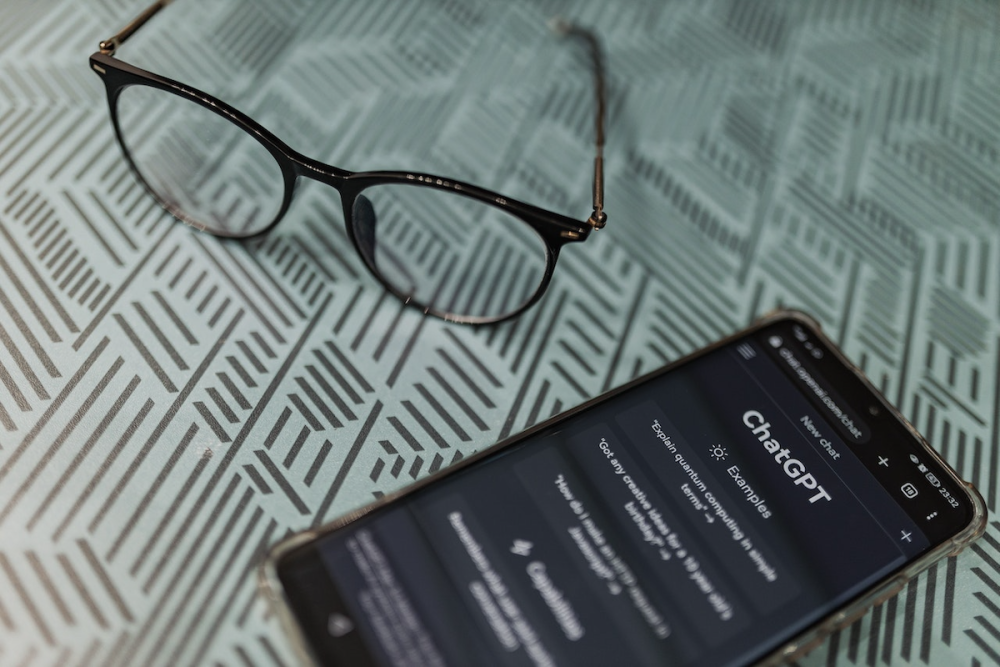 图片:一副眼镜放在智能手机旁边的桌子上，智能手机正在显示ChatGPT界面。
