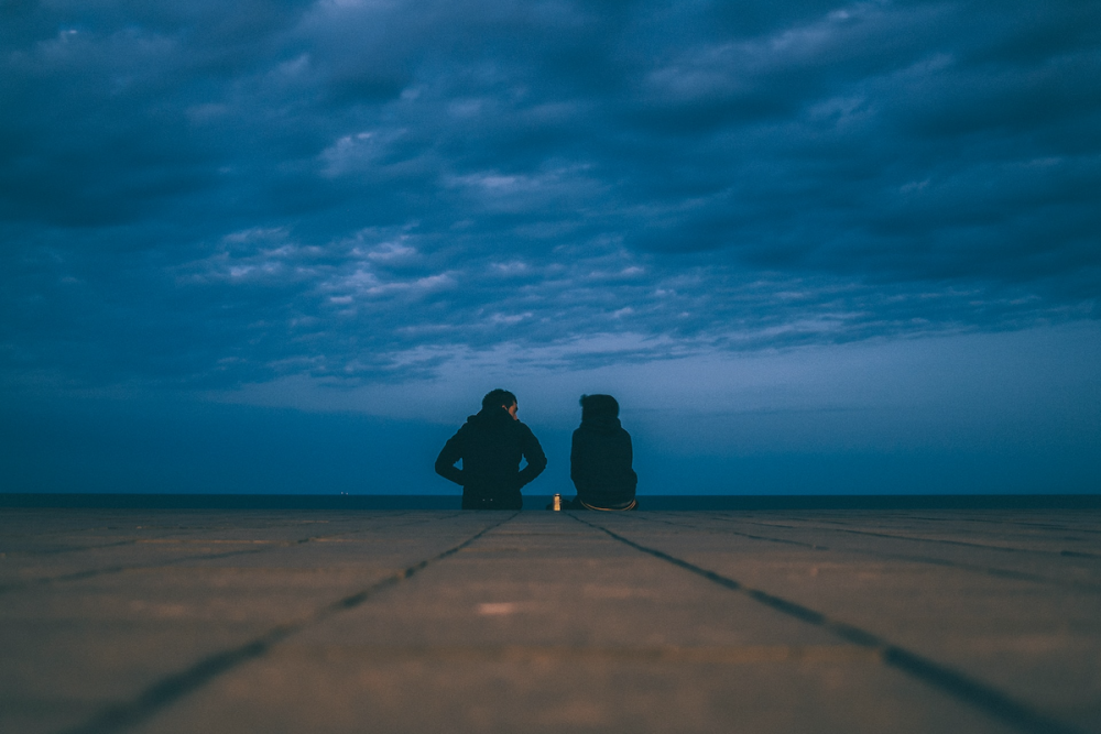 图片:从蠕虫的角度来看，一对穿着深色连帽衫的夫妇坐在砖墙的边缘，面对着开阔的海洋，在傍晚乌云密布的天空下。