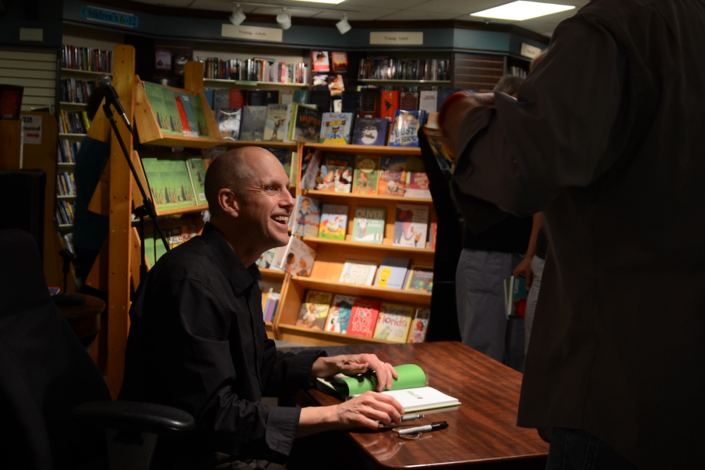 图片:一个作家微笑的观众在他准备亲笔签名的书在一个店内图书签售会。